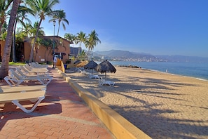 Un hôtel situé en bord de mer, doté de chaises longues et de parasols, offrant une vue dégagée sur l’océan et une chaîne de montagnes en arrière-plan.