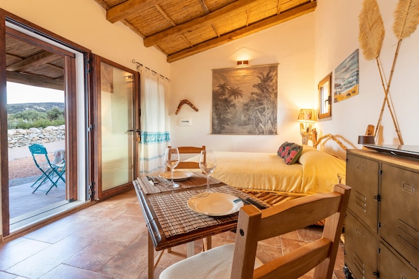 A room with a bed, a dining table with chairs, a view through a glass door, and a painting on the wall.