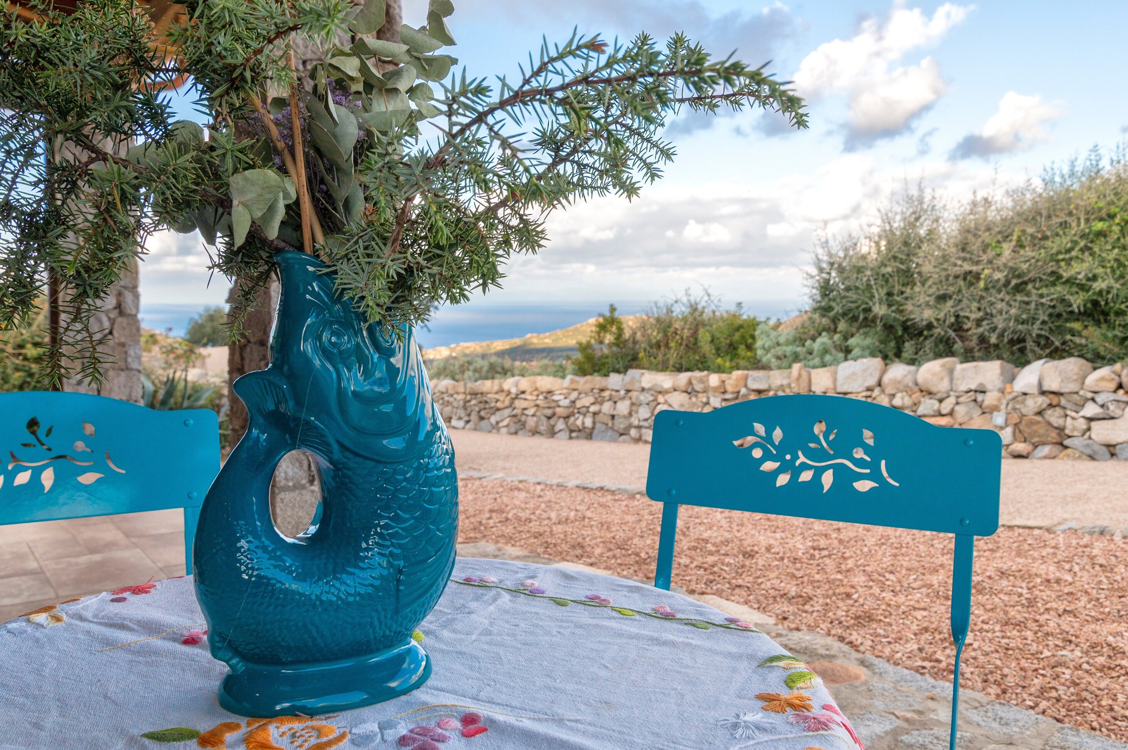 Ein Tisch mit einer blumigen Tischdecke, eine blaue Vase mit Grünpflanzen und eine Steinwand im Hintergrund.