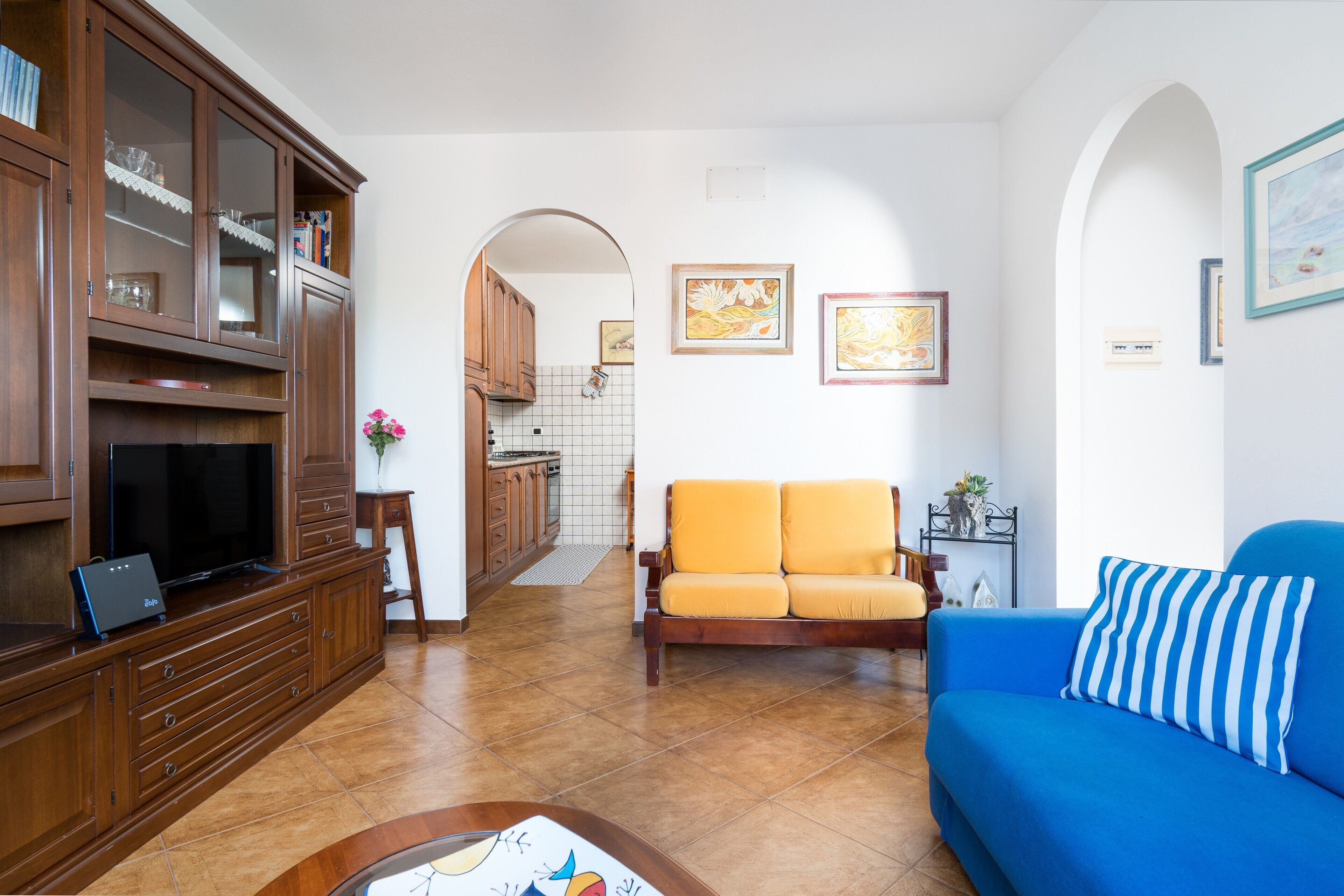 A living room with a blue sofa, a wooden entertainment unit, and a kitchen in the background.