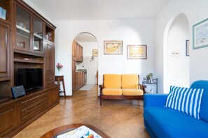 A living room with a blue sofa, a wooden entertainment unit, and a kitchen in the background.