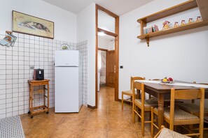 Une cuisine équipée d’un réfrigérateur, d’une table en bois, de chaises, d’un plan de travail carrelé, d’une étagère avec des objets décoratifs et d’un tableau encadré au mur.