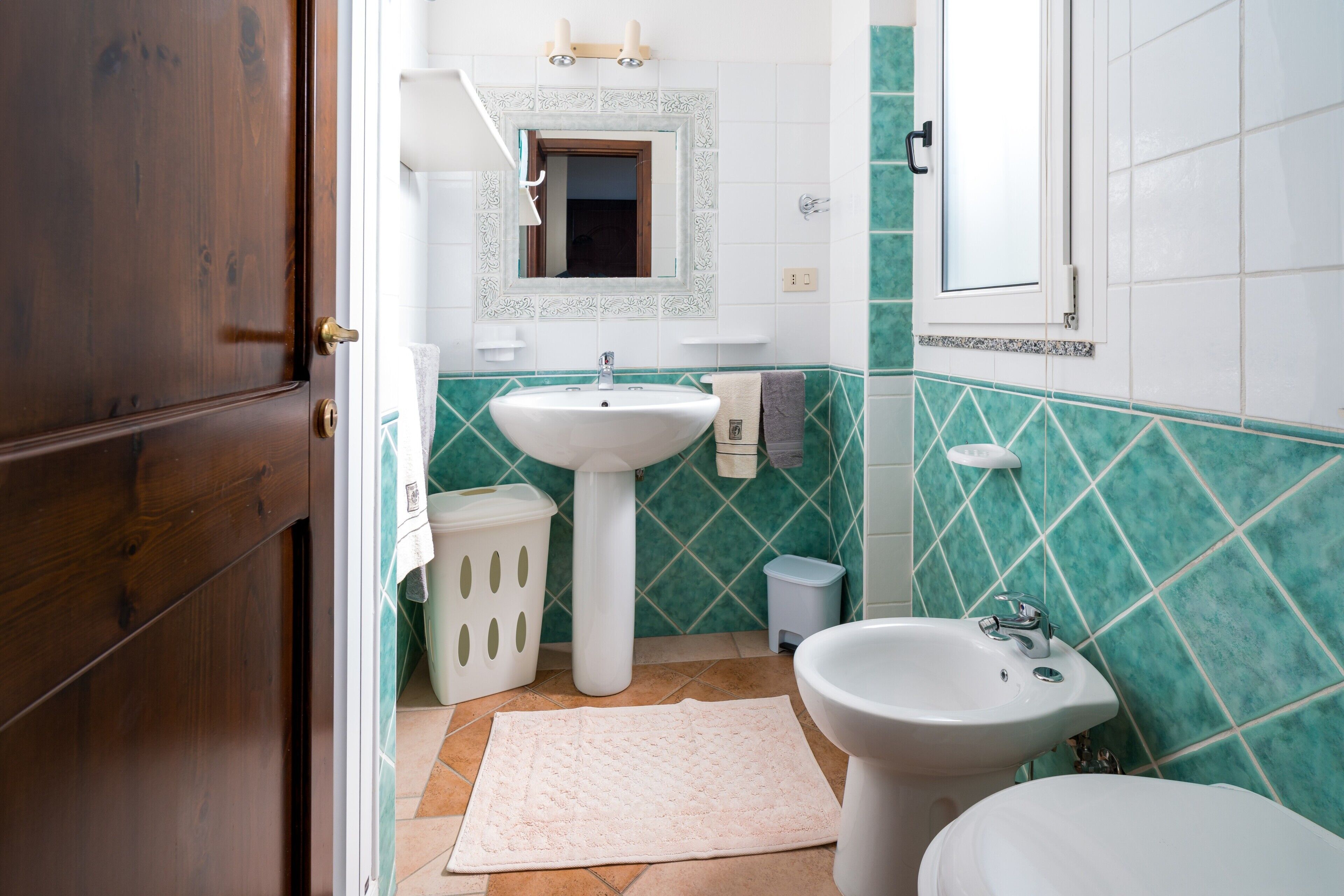 A bathroom with a wooden door, a pedestal sink, a bidet, a small trash bin, and a white towel on the floor.