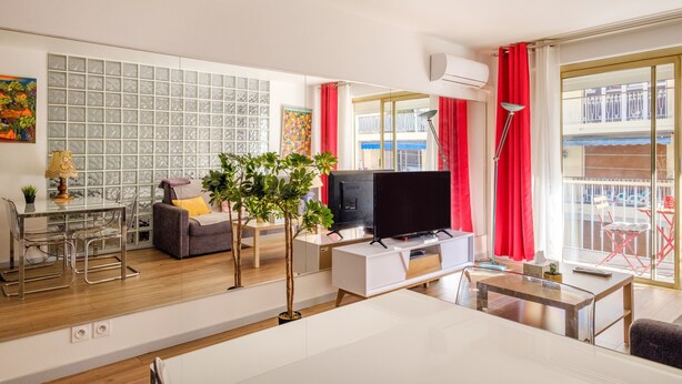 Una sala de estar moderna con televisor, sofá, mesa de comedor, sillas y un balcón con cortinas rojas.