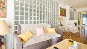 Una sala de estar con un sofá gris, una mesa de centro, una pared de bloques de vidrio y un comedor con mesa y sillas de vidrio.