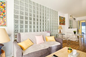 A living room with a grey sofa, a coffee table, a glass block wall, and a dining area with a glass table and chairs.