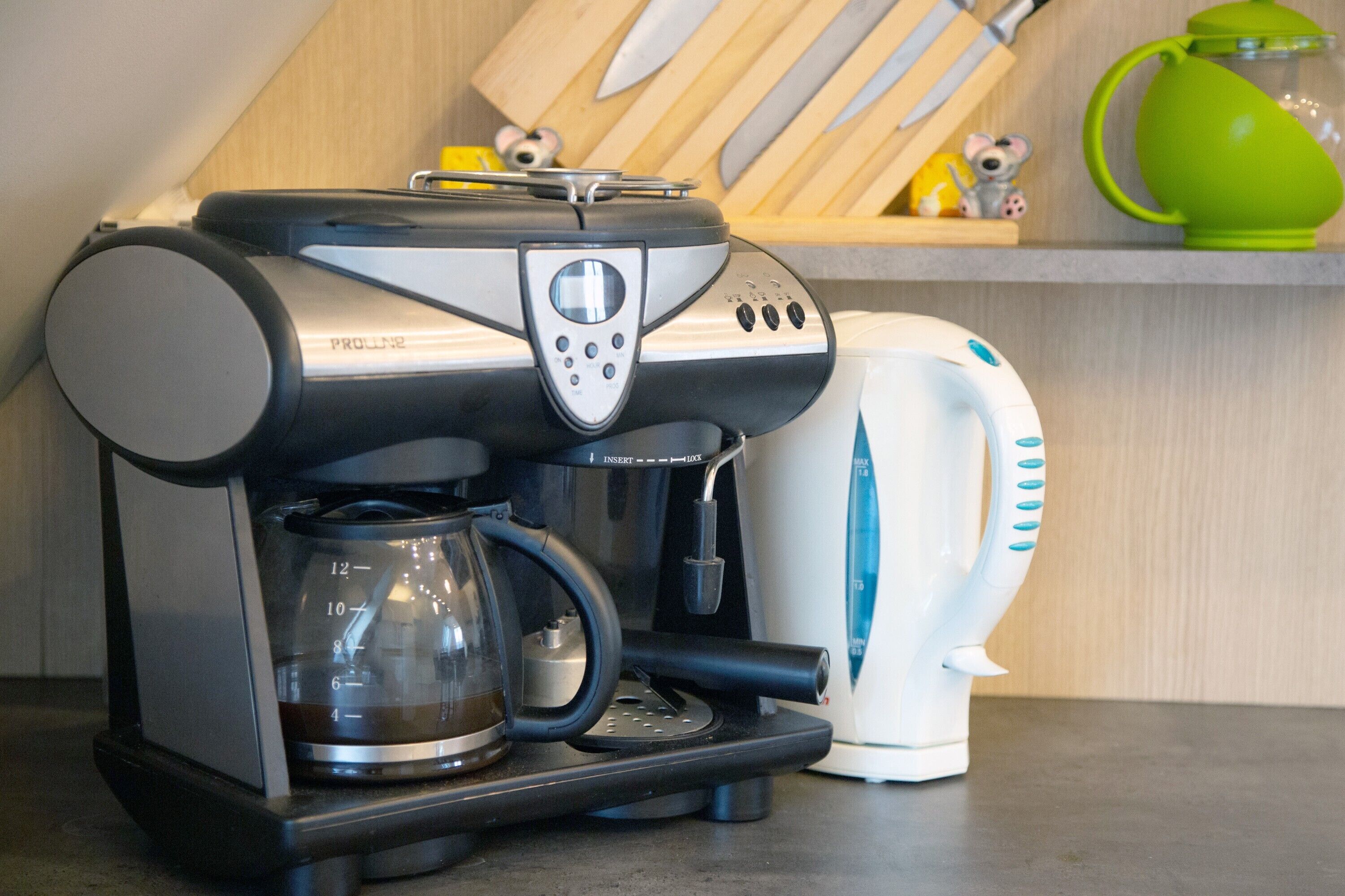 A coffee maker with a glass carafe, a white electric kettle, and a wooden shelf with kitchen utensils.