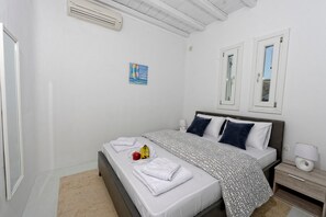 A neatly made bed with white linens, a patterned blanket, and a fruit bowl with bananas and an apple. There is a bedside table with a lamp and a small drawer. The room has white walls, a ceiling with exposed beams, and a window with a view. - Villa "Harmony' with Sea View, Private Pool & Wi-Fi (Mykonos)