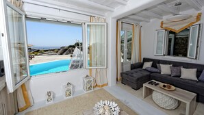A room with a view of a pool and beach, a grey sofa, a wooden coffee table, and a white decorative bowl. - Villa "Harmony' with Sea View, Private Pool & Wi-Fi (Mykonos)