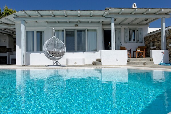 Una piscina con agua cristalina, una silla colgante y un edificio con pared de piedra y fachada blanca.