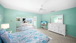 Ein Schlafzimmer mit Bett, Kommode, Schreibtisch und Fernseher. Ein Glastür führt auf einen Balkon mit Blick auf das Meer.