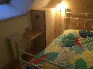 Ein kleiner, gemütlicher Raum mit schrägen Wänden, einem Holztisch, einem Metallbettgestell und einer farbenfrohen Bettdecke.