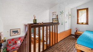Une pièce avec un escalier en bois, un lit recouvert d’une couverture bleue, un coin repas avec des chaises et une table, et un mur orné de tableaux.
