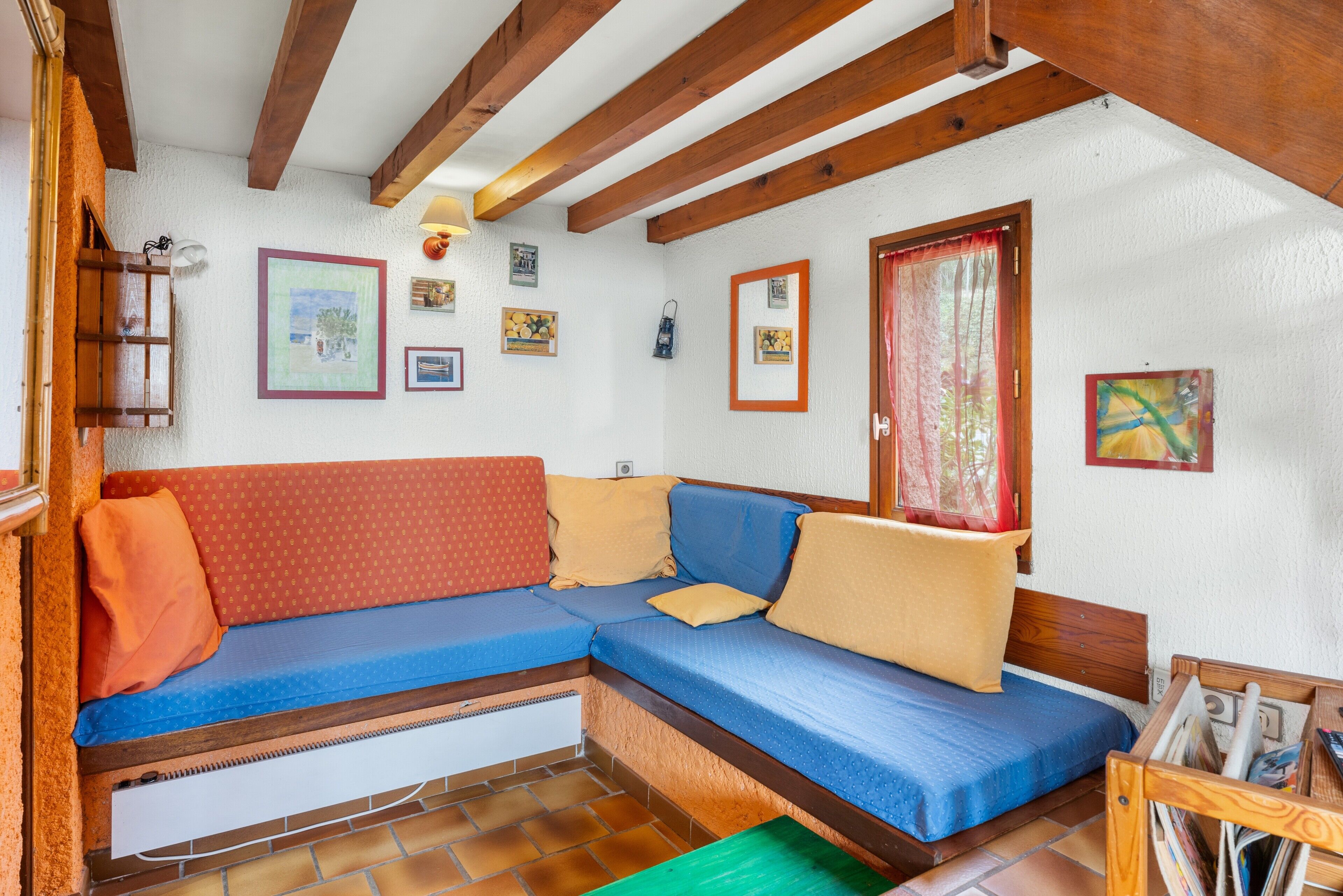 Une pièce chaleureuse avec un plafond en bois, un canapé bleu agrémenté de coussins orange et beige, une petite table avec des magazines et une fenêtre ornée d’un rideau rouge.