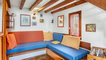 Une pièce chaleureuse avec un plafond en bois, un canapé bleu agrémenté de coussins orange et beige, une petite table avec des magazines et une fenêtre ornée d’un rideau rouge.