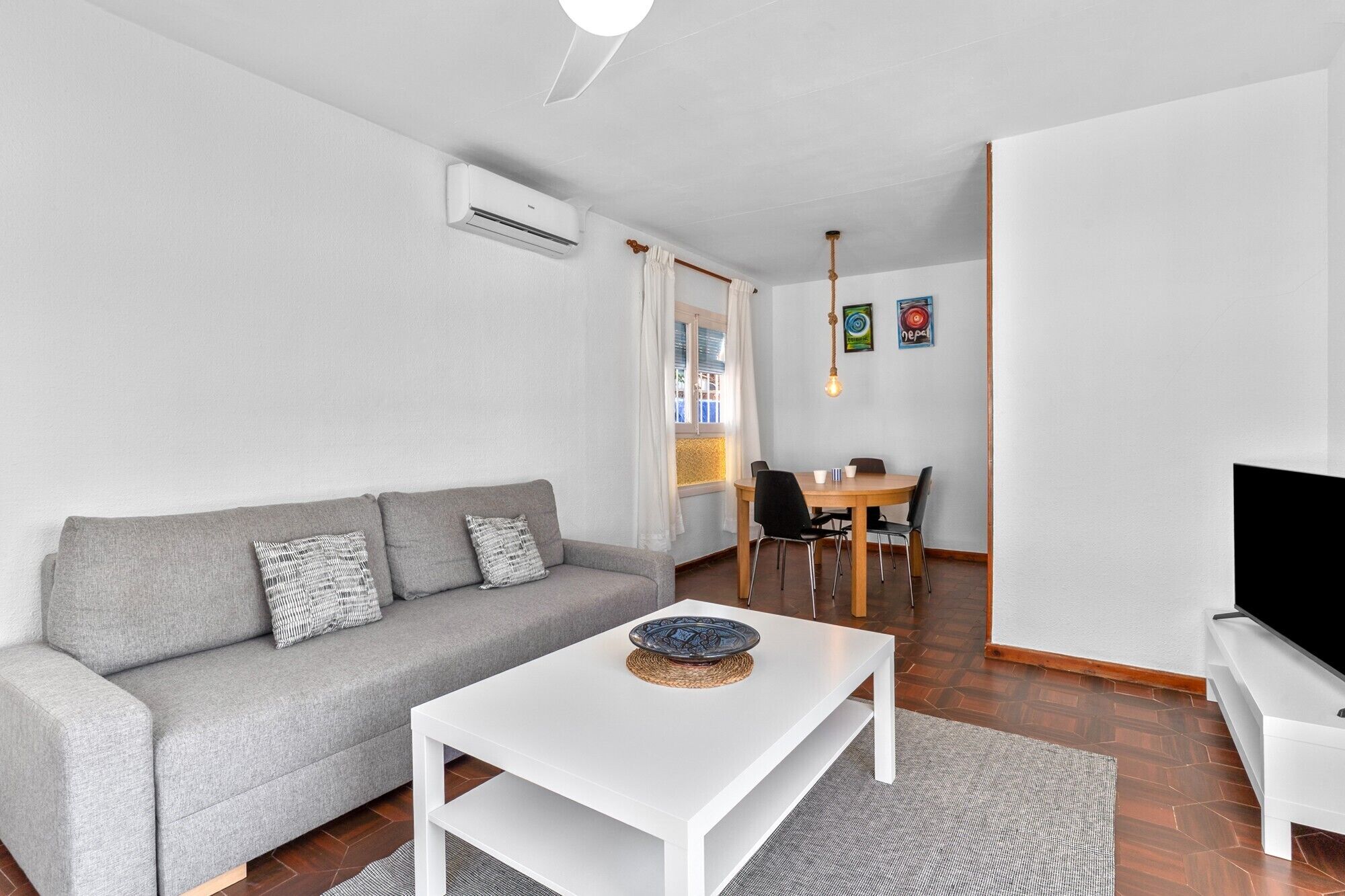 Una sala de estar moderna con un sofá gris, una mesa de centro blanca, un comedor con una mesa y sillas de madera, y un televisor sobre un mueble.
