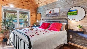 A bedroom with a wooden ceiling, a bed with red pillows, a white bedspread, and a balcony with a view of greenery. - Family Oasis By Ghosal Luxury Lodging (Sevierville)