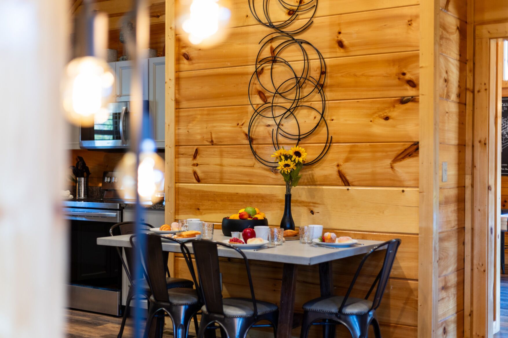 L’intérieur est en bois et comprend une table à manger dressée pour deux personnes, entourée de chaises. Un vase rempli de tournesols est posé sur la table, et on y trouve divers aliments et boissons.