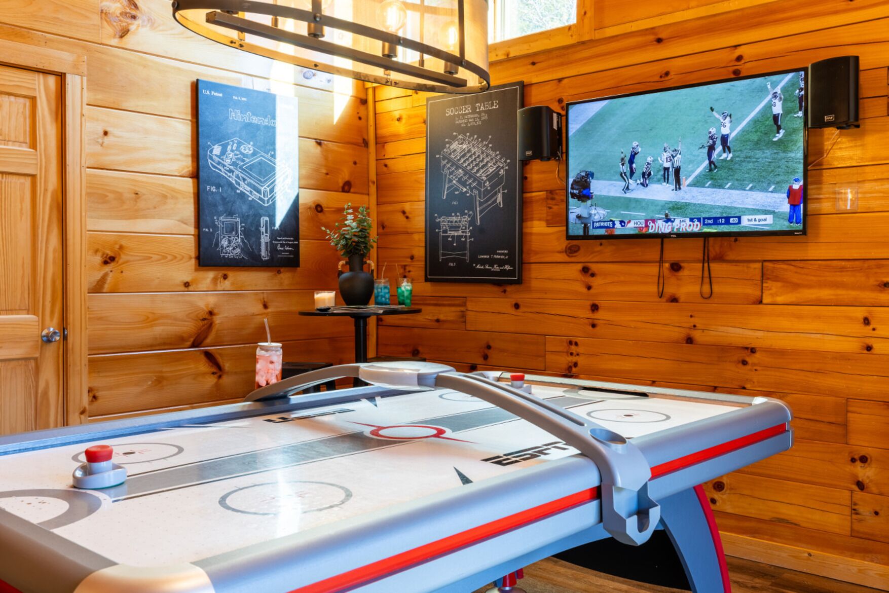 Un interior de madera con mesa de billar, un televisor mostrando un partido deportivo y carteles enmarcados en la pared.