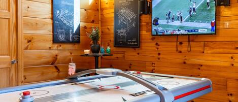Un interior de madera con mesa de billar, un televisor mostrando un partido deportivo y carteles enmarcados en la pared.