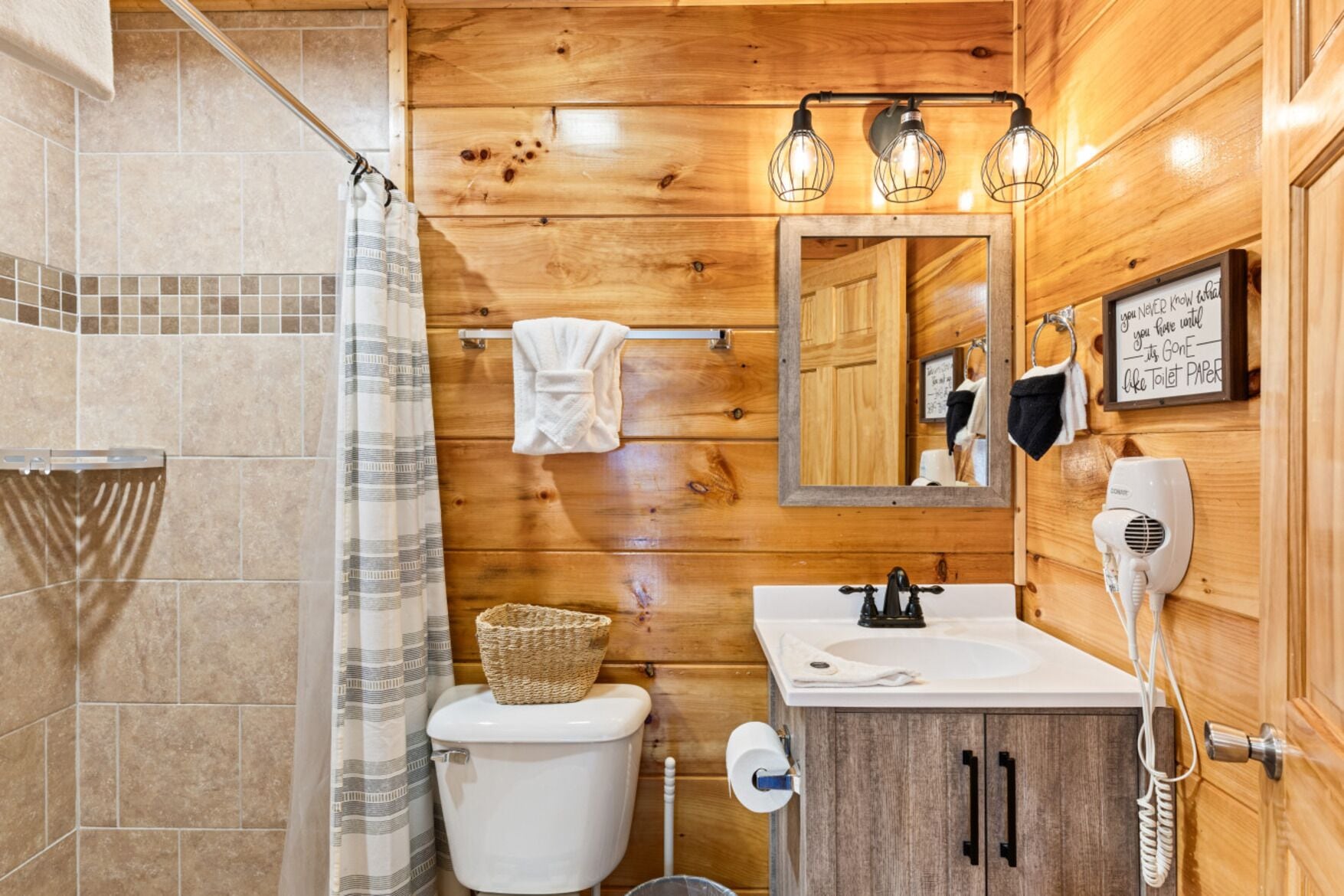 A wooden-paneled bathroom with a toilet, sink, and mirror. There are three wall-mounted lights, a hairdryer, and a towel rack.