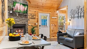 A cozy cabin interior with a stone fireplace, a dining table set with breakfast, a gray sofa, and a sign that reads "RELAX AND UNWIND". - Family Oasis By Ghosal Luxury Lodging (Sevierville)