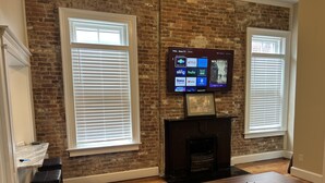 A living room with a brick wall, two large windows with blinds, a flat-screen TV mounted above a fireplace, and a wooden floor. - Historic Loft in Frankfort Entertainment District (Frankfort)