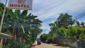 Placa indicando o Flower Garden Hostel, com uma estrada que leva até ele, rodeado por vegetação exuberante e um céu claro.
