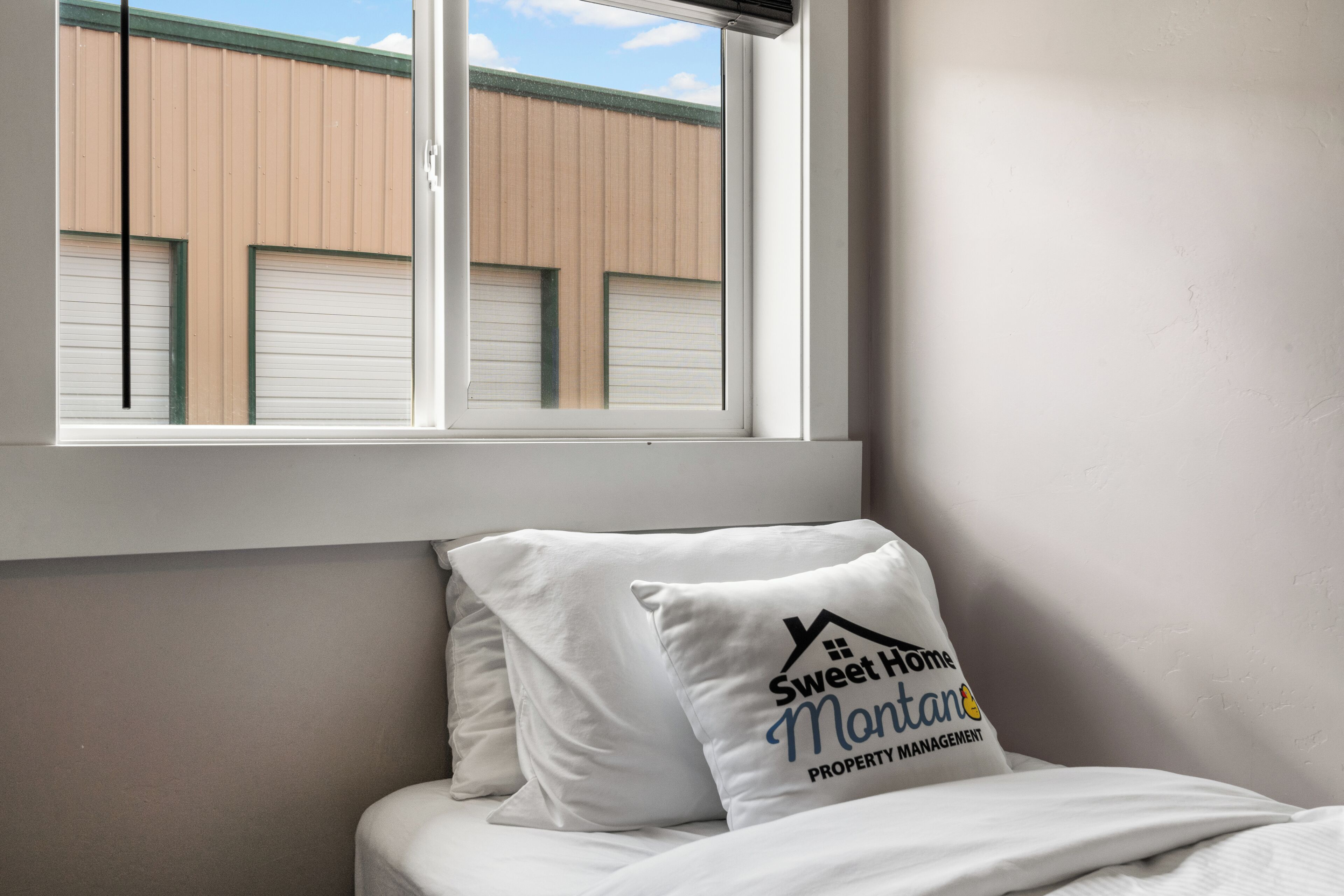 A bedroom with a window, a bed with white bedding, and a pillow with the text "Sweet Home Montana Property Management".