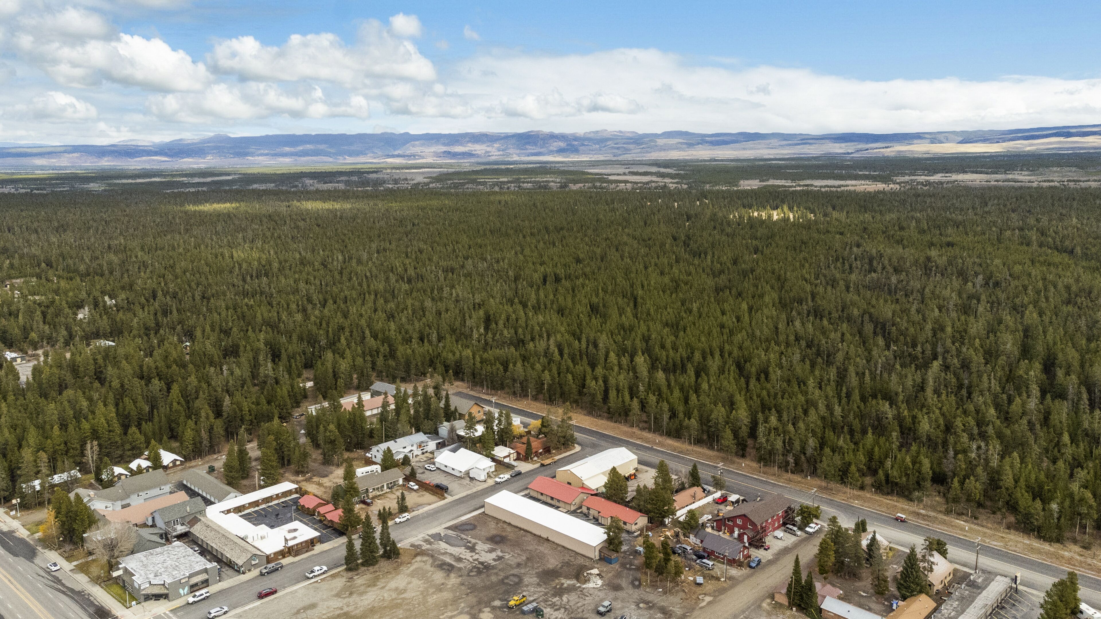 Ein Luftbild einer kleinen Stadt, umgeben von dichtem Wald, mit einer Hauptstraße und mehreren Gebäuden, darunter eine große Industrieanlage und Wohnhäuser.