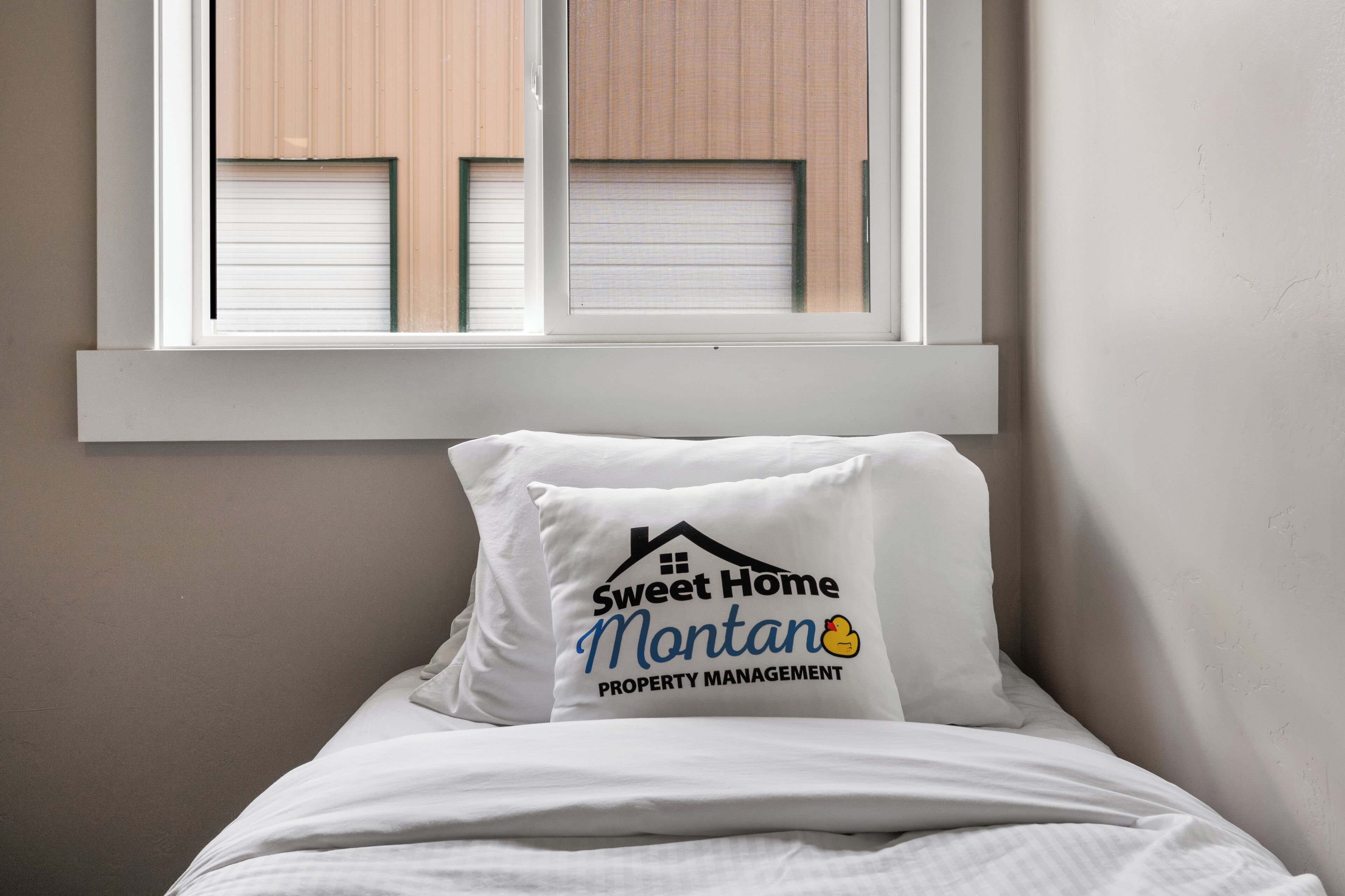 A bed with a pillow featuring a logo and text, a window with blinds, and a plain wall.