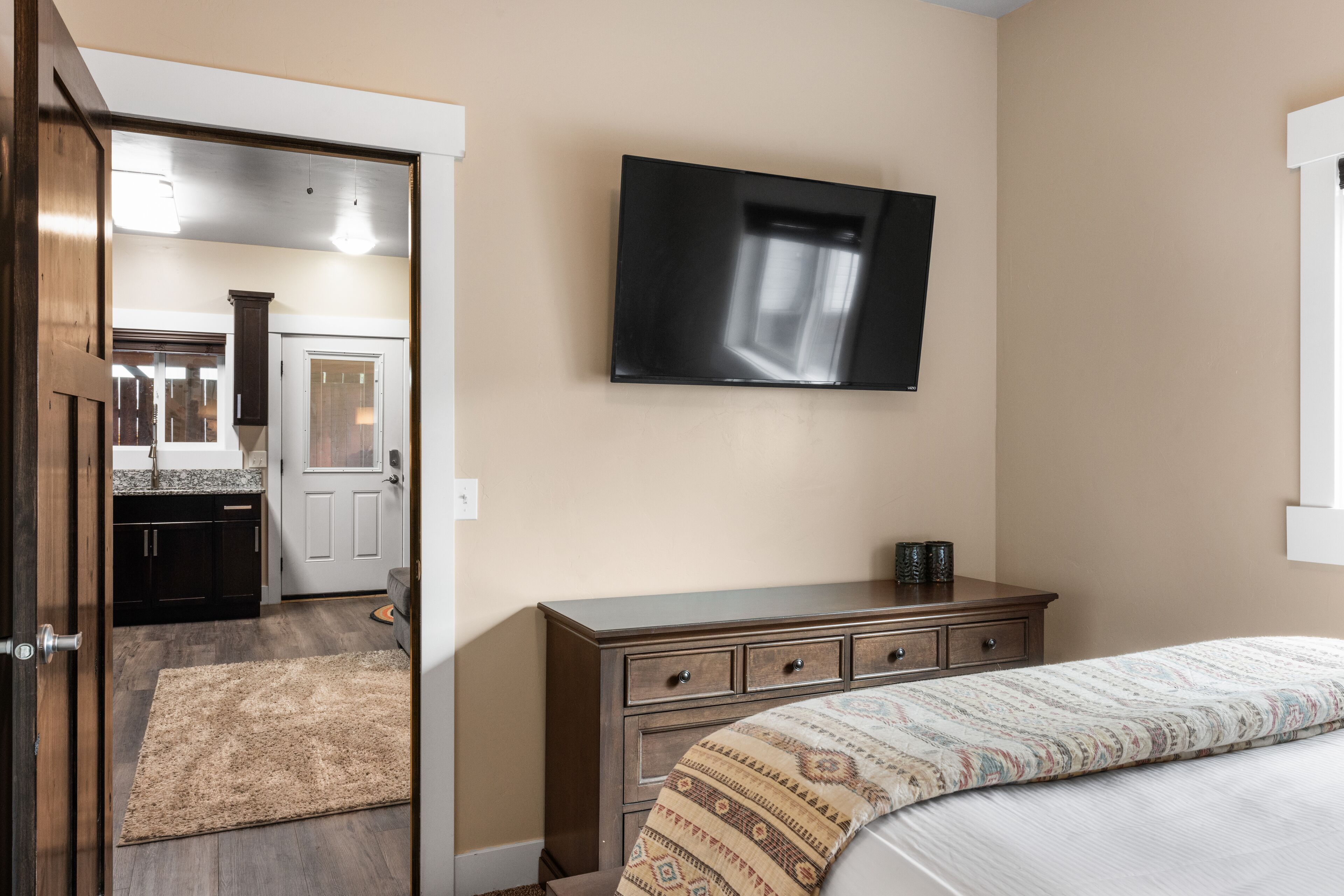 A bedroom with a bed, a dresser, a television mounted on the wall, and a door leading to a bathroom.