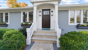 Une maison à deux étages avec un porche à l’avant, une porte marron et un petit jardin devant, agrémenté d’arbustes taillés et de jardinières.