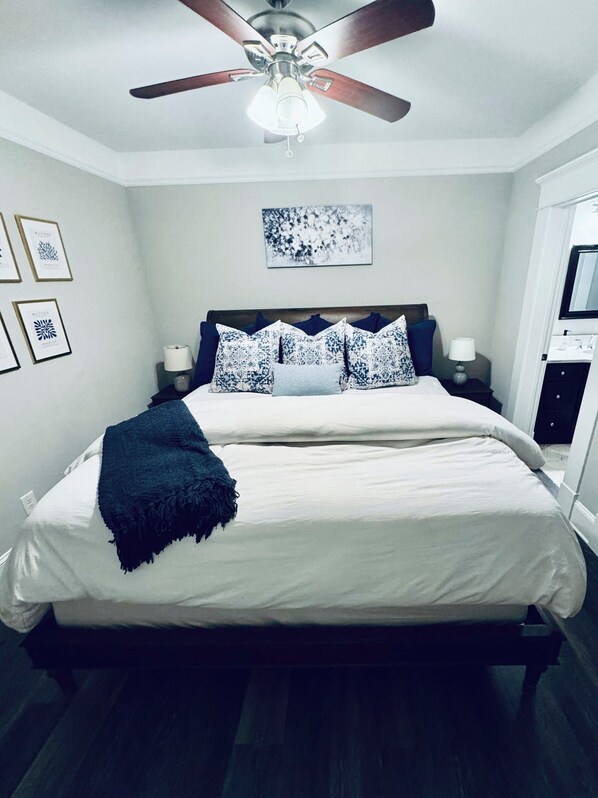 Un dormitorio con una cama grande, un ventilador de techo, obras de arte enmarcadas en la pared y una mesita de noche con lámpara.