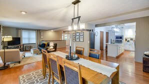 Ampla sala de estar com uma mesa de jantar de madeira, cadeiras e um lustre moderno. Cozinha visível ao fundo, com armários brancos e micro-ondas.