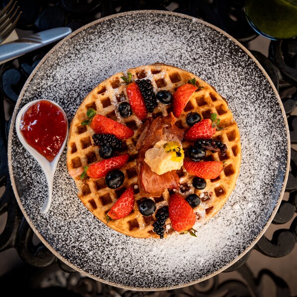 A waffle topped with strawberries, blueberries, blackberries, and a piece of bacon, served on a speckled plate with powdered sugar.