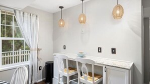 Un coin repas avec une table et des chaises blanches, trois lampes suspendues et une fenêtre avec des rideaux.