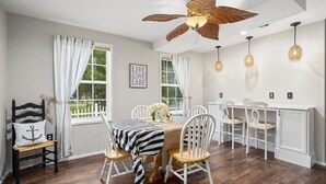 Un coin repas avec une table dressée pour quatre personnes, des chaises blanches, un ventilateur de plafond et une vue sur un lac par les fenêtres.