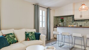 Een moderne woonkamer met een bank, twee witte bijzettafels en een keukenhoek met een barkrukset.