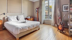 A bedroom with a large bed, wooden floor, a window with curtains, a guitar, and a bookshelf.