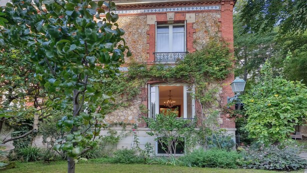 A two-story building with a brick facade, a balcony with wrought iron railings, and a garden area with various plants and a tree.