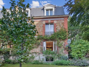 Ein zweistöckiges Gebäude mit Ziegelaußenwand, einem Balkon mit schmiedeeisernen Geländern und einem Gartenbereich mit verschiedenen Pflanzen und einem Baum.
