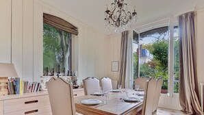 A dining room with a large wooden table set for six, surrounded by upholstered chairs. There is a chandelier above the table, and the room has large windows with curtains.