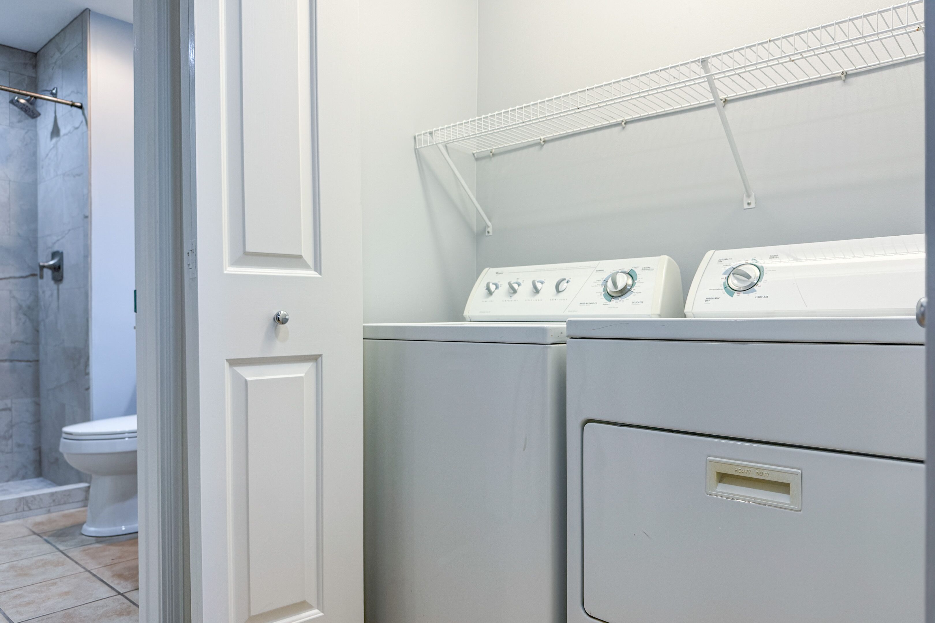 A laundry area with two washing machines, a metal shelf above them, and a toilet visible in the background.
