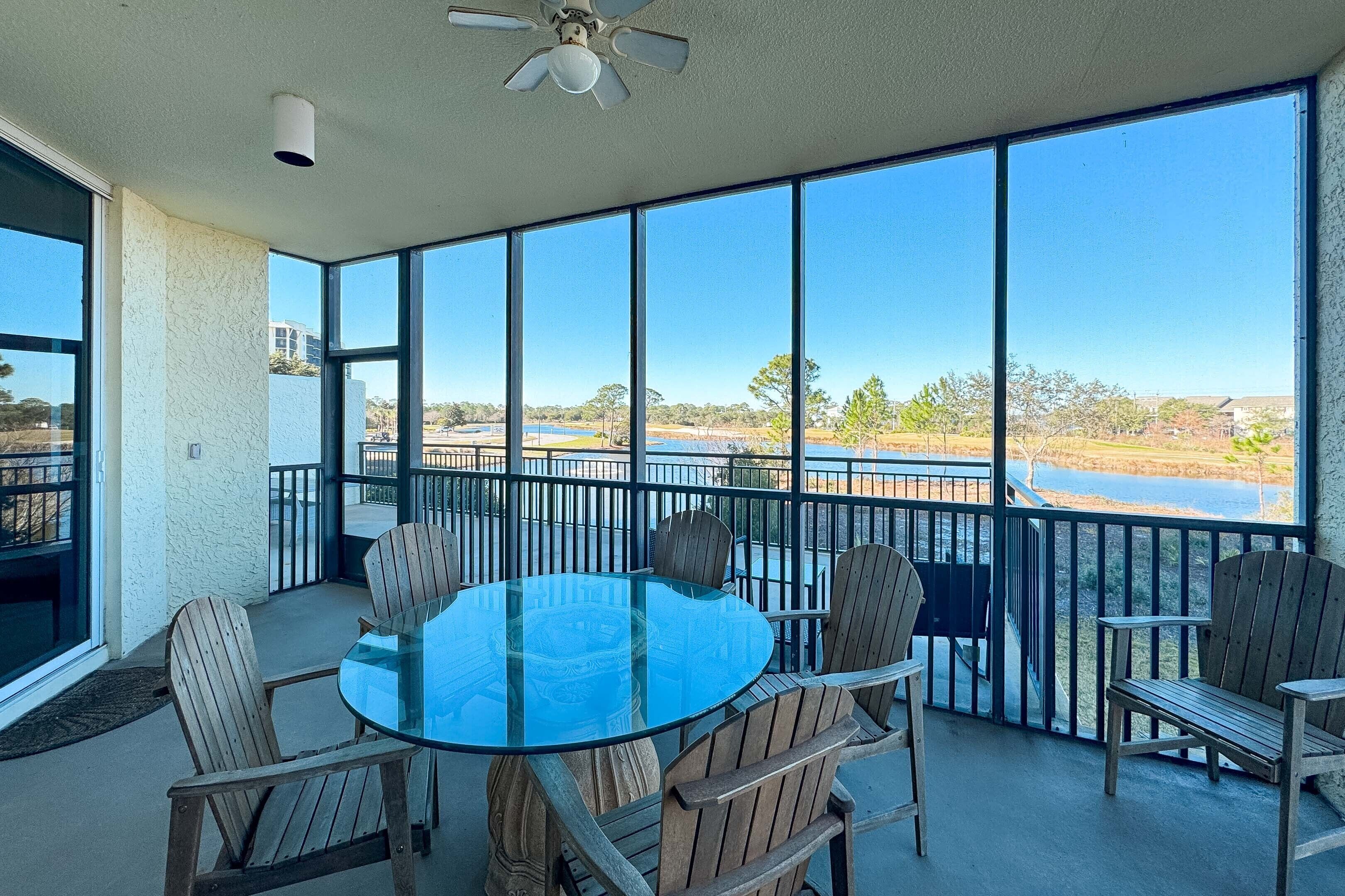 A glass-top table with wooden chairs on a balcony with large windows and a view of a lake.