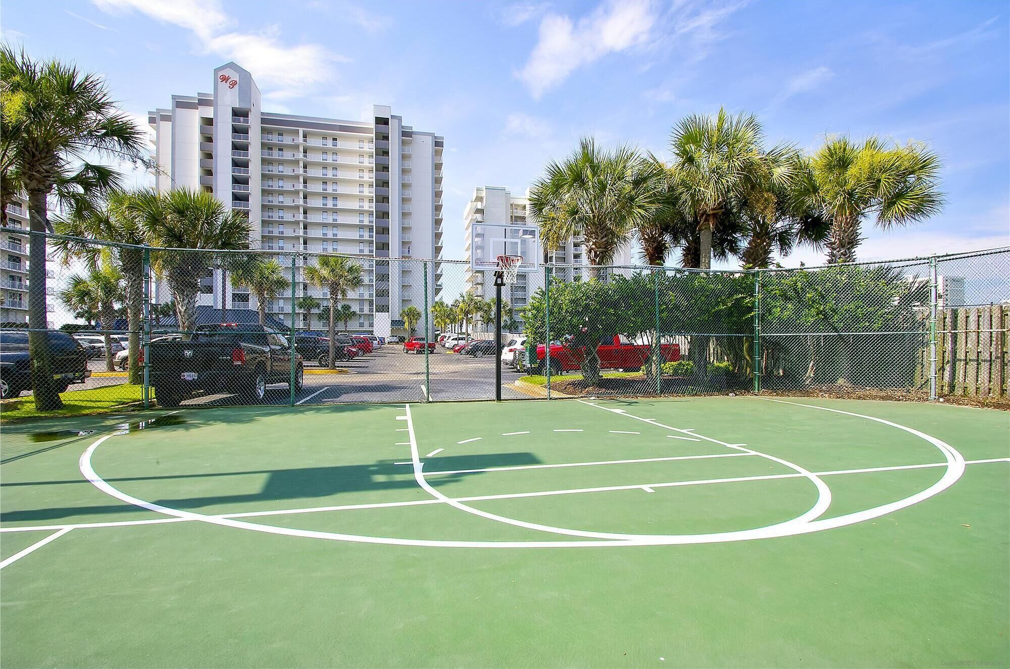 A basketball court with a chain-link fence, palm trees, and a multi-story building in the background.