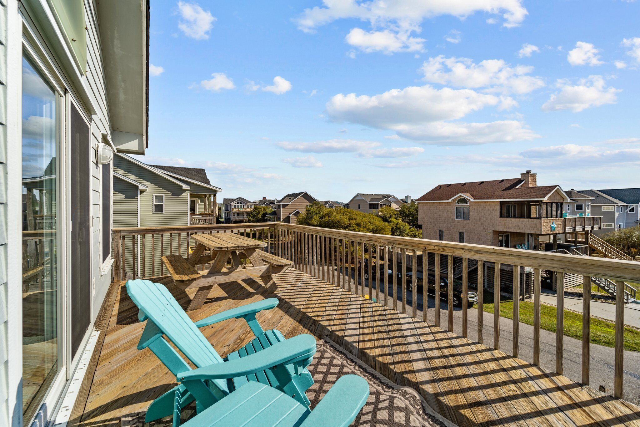 A wooden deck with blue Adirondack chairs, a small table, and a railing overlooking a residential area.
