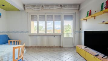 Una stanza con un letto, una TV, un termosifone, una finestra con tende e uno scaffale con oggetti.
