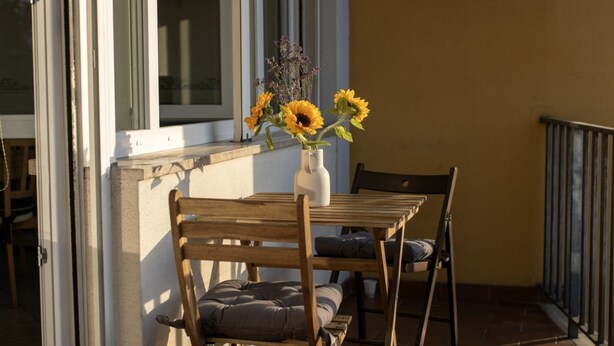 Ein Holztisch mit einer Vase voller Sonnenblumen und violetten Blumen, zwei Stühle mit Kissen und ein Fenster mit Blick ins Freie.