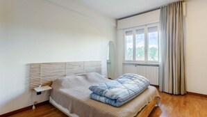 Ein Schlafzimmer mit Bett, Nachttisch, Fenster mit Vorhängen und Blick auf Grünfläche.